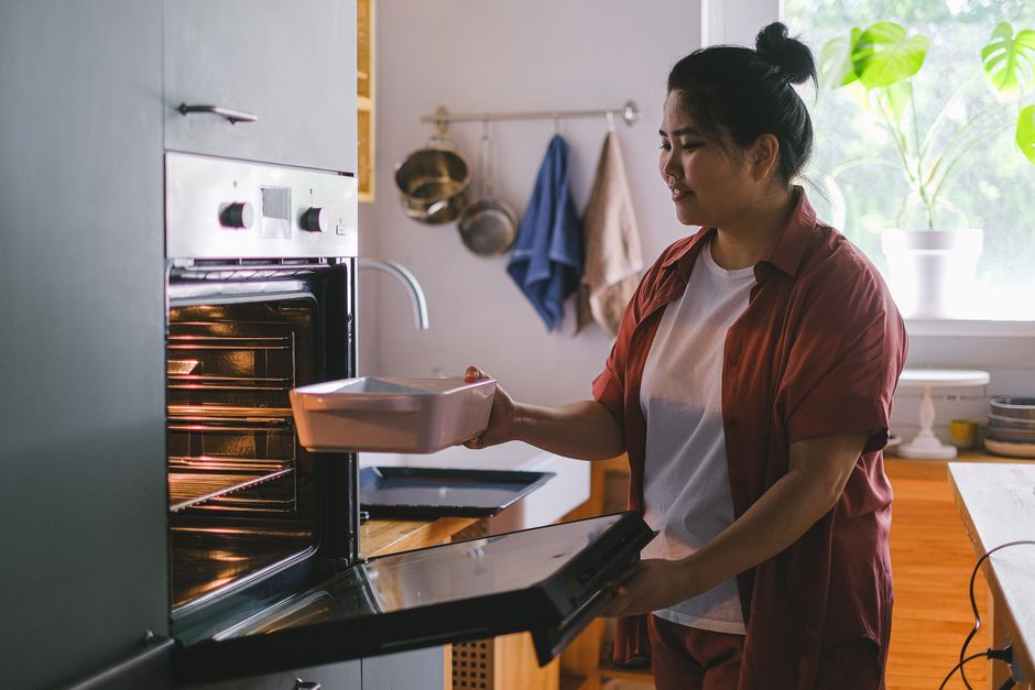 Close-up of a black, ceramic oven surface being cleaned during a domestic deep cleaning process by [COMPANY_NAME], with a person wearing blue rubber gloves wiping the oven's interior surface with a mi